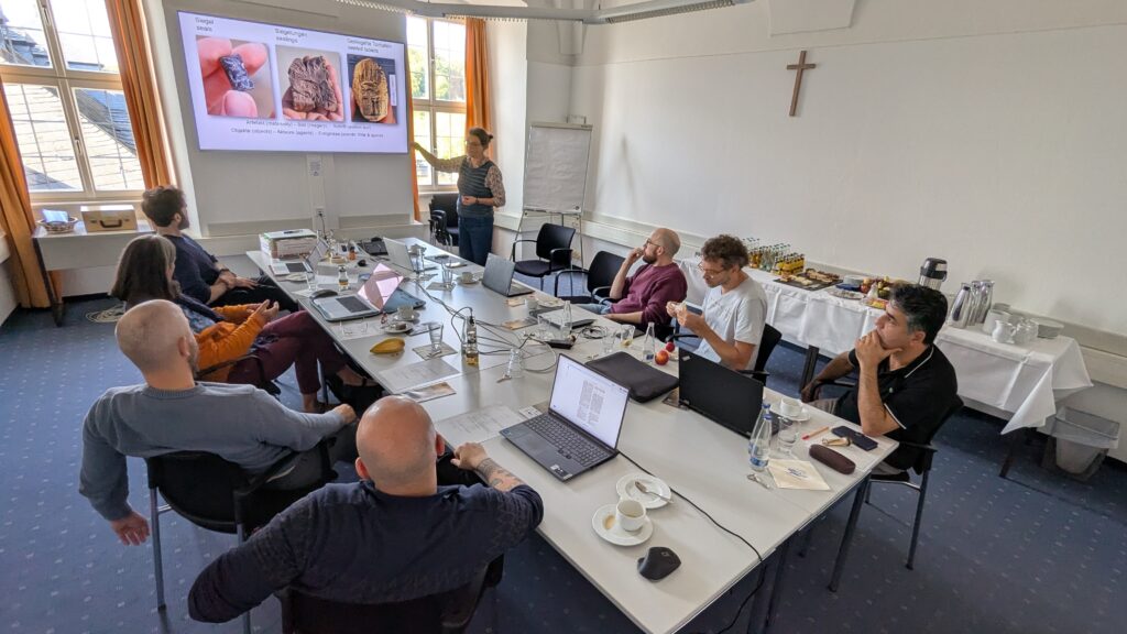A group of people sitting at a conference table. One person is giving a presentation. There are laptops and notes on the table and a small buffet in the background.