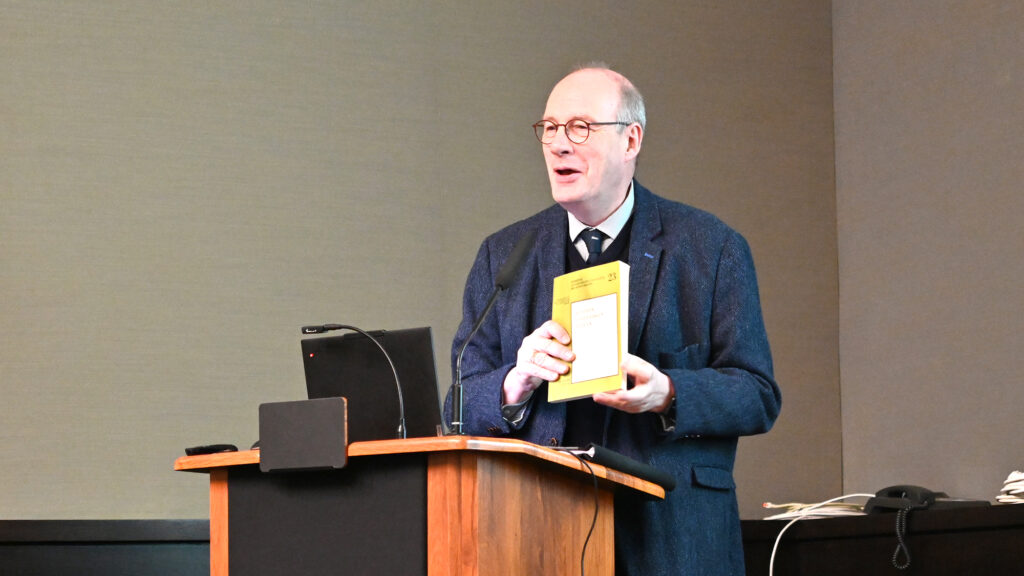 A person behind a podium holding a book and talking to an invisible audience.