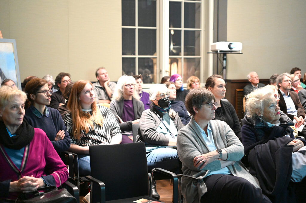 Half a room full of people listening to an invisible speaker. A window in the background.