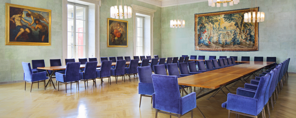 Ein Raum mit Tischen in U-Form und blauen Stühlen. An der Wand hängen zwei Gemälde und ein Wandteppich.