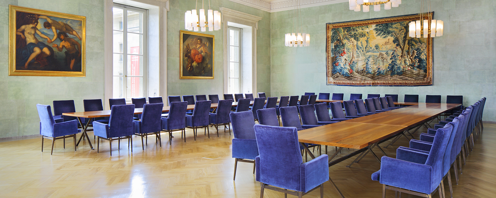A room with tables shaped in u-form and blue chairs. The wall depicts two paintings and a wall carpet.